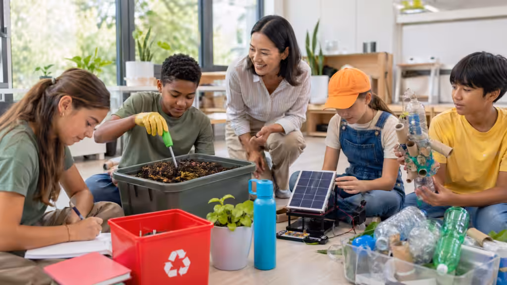 4-5 diverse K-12 students working together on sustainability hands-on projects: compost, solar panel kit, recycled materials sculpture; teacher kneeling beside them